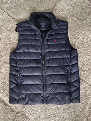 Mörkblå dunväst från Polo Ralph Lauren - Snygg mörkblå dunväst från Polo Ralph Lauren med quiltad design och hög krage. Västen har två dragkedjeförsedda fickor och en liten röd logga på bröstet. 