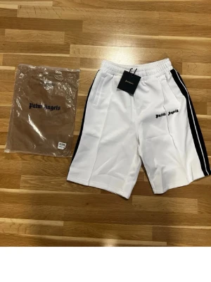 Vita Palm Angels shorts med svarta ränder - Palm Angels vita shorts med svarta sidoränder och broderad logga på vänster ben. Elastisk midja och sidofickor. Sportig och clean look, perfekt för sommaren.