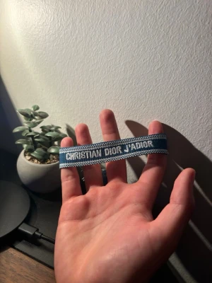 Dior armband - Snyggt blått tygarmband från Christian Dior med texten 'J'ADIOR' broderat i vitt. Armbandet har justerbara tofsar i ändarna och en cool, vävd design som ger en trendig vibe. Perfekt accessoar för att lyfta din outfit.
