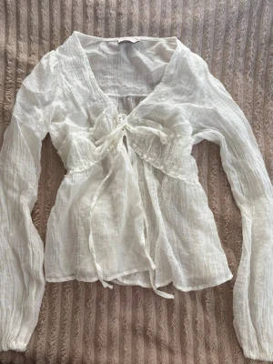 Vit omlottblus med knyt och volang - Säljer en superfin vit omlottblus i tunn, luftig bomull med långa ärmar och volangdetaljer. Blusen har v-ringning och knyt framtill som ger en romantisk vibe. Perfekt till shorts eller kjol för en fräsch look.