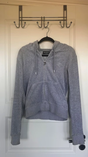 Grå zip hoodie från Juicy Couture - Säljer en grå zip hoodie från Juicy Couture med huva och dragsko. Hoodien har fickor framtill och ett broderat JC-logo på bröstet. Tillverkad i mjukt material som känns skönt mot huden. Perfekt för en chill och avslappnad stil. 