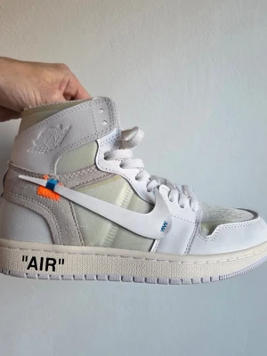 Air Jorden 1 Retro off white - Unik par vita high-top sneakers från Air Jorden 1 retro med genomskinliga paneler, vit sula och texten 'AIR' på sidan. Skorna har en vit swoosh med blå och orange sömdetalj samt präglad logga på ankeln. Materialmix av läder och mesh för en modern look.
