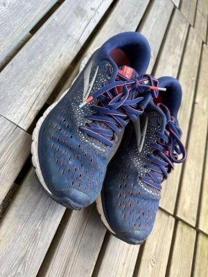 Blå Brooks sneakers med röda detaljer - Snygga sneakers från Brooks i marinblått med röda och vita detaljer. Ovandelen är i mesh för bra ventilation och skorna har vita, dämpade sulor. Perfekta för dig som gillar sportig stil och vill ha bekväma skor till vardags.