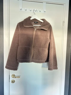 Brun teddyjacka med dragkedja - Mysig brun teddyjacka med hög krage och dragkedja framtill. Jackan har två stora fickor och en loose passform som ger en avslappnad look. Perfekt för kyliga dagar när du vill vara både varm och stilren.