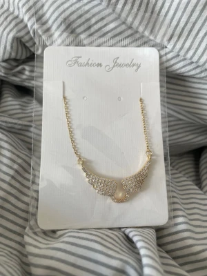 Guldhalsband med  vingar - Snyggt halsband i guldfärg med ett hängsmycke format som två vingar täckta av små glittrande stenar. Kedjan är tunn och elegant, perfekt för att ge din outfit lite extra bling. Passar dig som gillar detaljer och vill sticka ut.