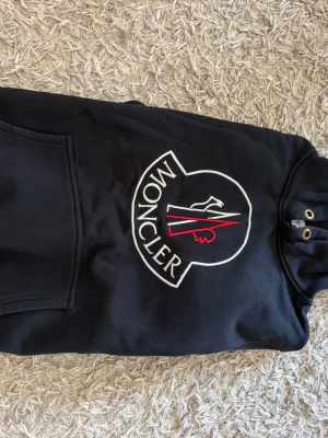 Svart Moncler hoodie med stor logga - Säljer en svart hoodie från Moncler med stor broderad logga i vitt och rött. Väldigt snygg och även väldigt skön och ha på sig tveka inte om det är några funderingar priset är inte hugget i sten