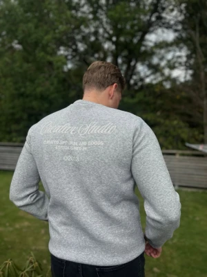 Grå stickad sweatshirt  - Ljusgrå sweatshirt från Creative Studio med broderad text på bröstet och större tryck på ryggen. Tröjan har rund hals, långa ärmar och ribbade muddar vid ärmslut och nederkant. Perfekt för en avslappnad och stilren look.