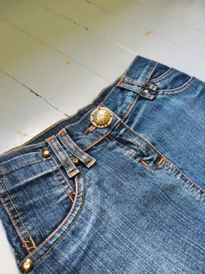 Blå jeanskjol - Säljer en blå jeanskjol. Kjolen har dekorativa nitar på fickorna och kontrastsömmar i orange. Klassisk femficksmodell i denim med coola detaljer. Vet inte exakt storlek men skulle gissa på storlek 146.