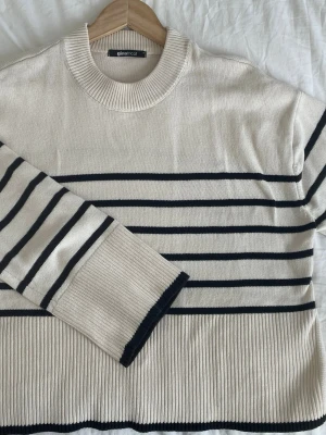 Randig stickad tröja från Gina Tricot - Snygg stickad tröja från Gina Tricot i offwhite med svarta horisontella ränder. Tröjan har rund halsringning, långa ärmar och bred ribbstickad kant nedtill och vid ärmsluten. Perfekt för en trendig och avslappnad look.
