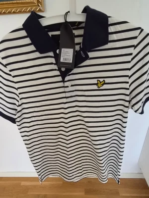 Oanvänd Randig piké från Lyle & Scott - Säljer en snygg vit och marinblå randig pikétröja från Lyle & Scott. Tröjan har korta ärmar, klassisk krage och knappslå framtill. Tillverkad i mjuk bomull och har den ikoniska gula loggan broderad på bröstet.