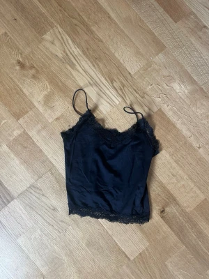Svart linnetopp med spetsdetaljer - Snygg svart linnetopp med tunna axelband och spetsdetaljer längs halsringning och nederkant. Tillverkad i ett mjukt, glansigt material som ger en lyxig känsla. Perfekt att styla lager på lager eller bära som den är.