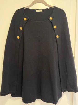 Stenströms Mary svart merinoull cape - Snygg klassisk svart stickad cape från Stenströms med sex dekorativa guldknappar framtill. Perfekt för dig som vill ha en unik och trendig look. Materialet ser ut att vara mjukt och bekvämt.