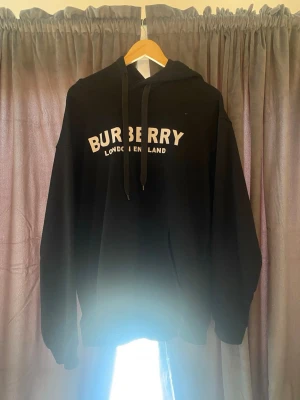Svart Burberry hoodie med tryck - Säljer en svart hoodie från Burberry med vit text 'BURBERRY LONDON ENGLAND' på bröstet. Klassisk modell med huva och dragsnören, samt känguruficka framtill. Perfekt för dig som gillar stilrena och exklusiva plagg.
