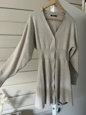 Beige kort stickad klänning Gina Tricot - Kort stickad klänning från Gina Tricot i ett finstickat material. Den är beige och har använts enstaka gånger. Klänningen har v-ringning, långa ärmar och knappar hela vägen framtill.