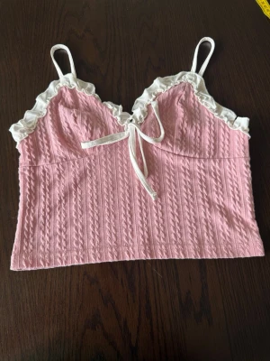 Rosa linnetopp med volang och knyt - Supersöt rosa linnetopp med vita volangdetaljer och knyt framtill. Toppen har smala axelband och är ribbad med ett mjukt, stretchigt material. Perfekt för en trendig och feminin look.
