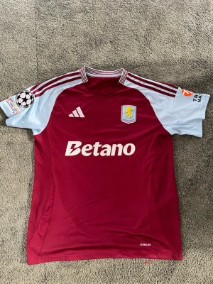 Aston Villa matchtröja vinröd/blå - Säljer en officiell Aston Villa matchtröja från Adidas i vinrött med ljusblå ärmar. Tröjan har klubbmärke, Betano-logga och Champions League-märke på ärmen. Tillverkad i lätt och ventilerande material, perfekt för fotbollsträning eller match.