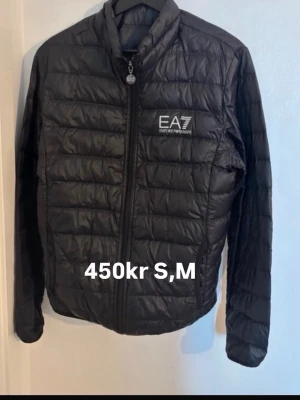 Svart pufferjacka från EA7 Emporio Armani - Snygg svart pufferjacka från EA7 Emporio Armani med diskret logga på bröstet. Jackan har quiltad design, dragkedja framtill och hög krage. Perfekt för kalla dagar och ger en stilren look. Tillverkad i lätt och värmande material.