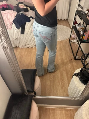 Ljusblå bootcut jeans med slitningar - Säljer ett par ljusblå bootcut jeans med slitningar på knäna och broderad detalj på bakfickan. Jeansen har klassisk femficksmodell och är tillverkade i denim med en skön passform. Perfekta för en avslappnad och trendig look.