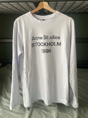 Vit långärmad t-shirt Acne Studios - Priset är EJ hugget i sten Vit långärmad topp från Acne Studios med tryck 'Acne Studios STOCKHOLM 1996' på bröstet. Klassisk rund halsringning och relaxed fit. Perfekt för dig som gillar minimalistisk och stilren design.