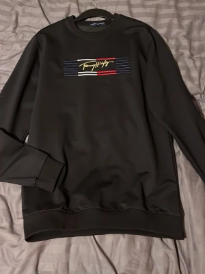 Svart sweatshirt Tommy Hilfiger - Svart sweatshirt från Tommy Hilfiger med broderad logga i gult, vitt och rött på bröstet samt en mindre logga på ärmen. Tröjan har rund halsringning, ribbade muddar och en stilren design som passar perfekt till jeans eller joggers.