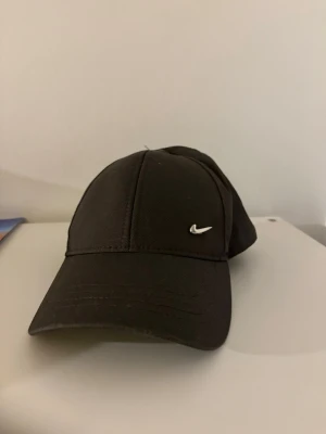 Svart keps från Nike  - Snygg svart keps från Nike med klassisk böjd skärm och den ikoniska vita swoosh-loggan broderad på framsidan. Tillverkad i slitstarkt tyg och justerbar baktill för perfekt passform. Enkel och stilren design som passar till det mesta. 
