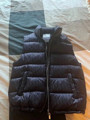 Blå/ lila väst  - Snygg mörkblå/ lila väst med diskret monogrammönster över hela västen. Västen har hög krage, dragkedja framtill och två fickor på sidorna. Perfekt för lager på lager och ger en lyxig vibe till din outfit. Endast testat på mig själv iprincip helt ny bara att de fattas prislapp på den. Pris kan diskuteras vid smidig affär