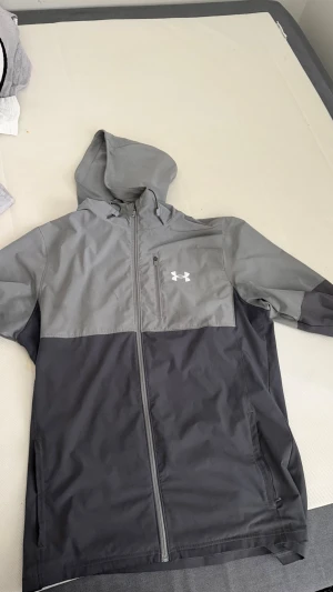 Grå vindjacka från Under Armour - Snygg tvåfärgad vindjacka från Under Armour i ljus- och mörkgrått. Jackan har huva, hel dragkedja framtill och en bröstficka med dragkedja. Tillverkad i lätt och vindtätt material, perfekt för blåsiga dagar.