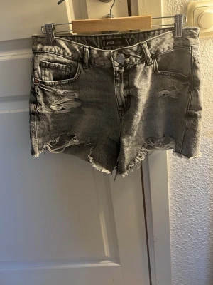 Grå slitna jeansshorts från FB Sister - Säljer ett par grå jeansshorts från FB Sister i storlek M. Shortsen har hög midja, råa kanter och slitna detaljer framtill för en edgy look. Perfekta till sommaren och lätta att matcha med olika toppar.