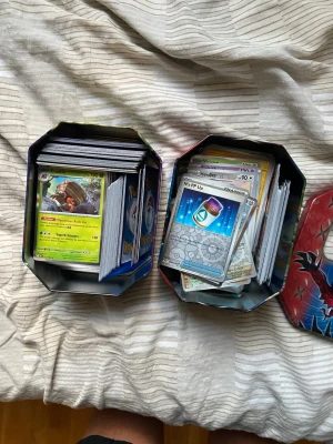 Pokémonkort i plåtlådor - En samling Pokémonkort i olika färger och motiv, förvarade i två plåtlådor med Pokémon-design. Korten är gjorda av papper och plast och innehåller både tränarkort, energikort och vanliga Pokémonkort. Perfekt för dig som gillar samlarkort och Pokémon.