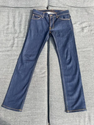 Mörkblå raka jeans med kontrastsömmar - Helt nya mörkblå Nudie jeans med raka ben och klassiska kontrastsömmar. Midja: ca 38,5cm, benlängd: ca 97cm Jeansen har fem fickor och dekorativa sömmar på bakfickorna. Tillverkade i denim med en stilren look och patch i läderimitation bak.