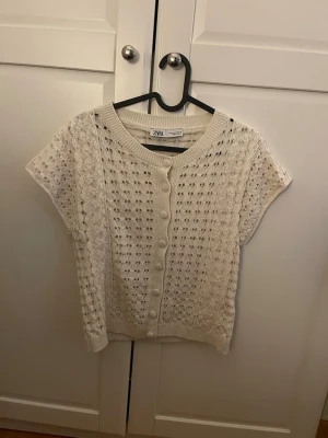 Vit kortärmad stickad kofta Zara - Söt vit stickad kofta från Zara med korta ärmar och rund hals. Koftan har dekorativa knappar framtill och ett hålmönster över hela plagget. 