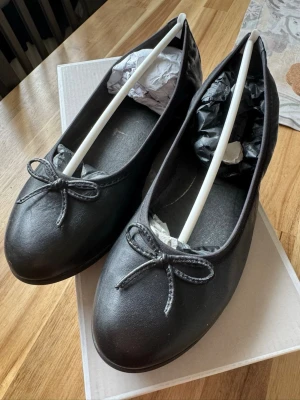 Svarta klassiska ballerinaskor - Stilrena svarta ballerinaskor med rund tå och dekorativ rosett framtill. Skorna har en enkel och tidlös design i matt material som passar till många olika outfits. Perfekta för dig som gillar en minimalistisk look.