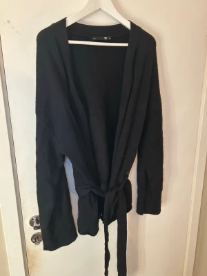Svart cardigan med knytskärp XL - Svart långärmad cardigan från 157 i storlek XL. Modellen är öppen framtill och har ett matchande knytskärp i midjan. Materialet ser ut att vara mjukt och stickat, perfekt för lager på lager och chill stil.