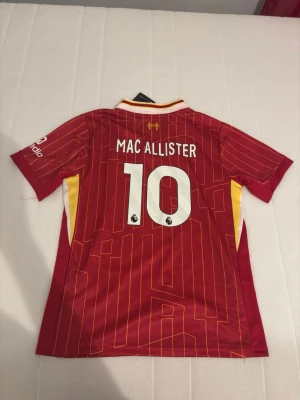 Liverpool matchtröja Mac Allister #10 - Röd Liverpool matchtröja med gula detaljer och vita ränder. Tryck med Mac Allister och nummer 10 på ryggen. Nike-logga och Liverpool FC-märke på bröstet, samt sponsortryck. Tillverkad i lätt och ventilerande material, perfekt för fotboll eller träning.