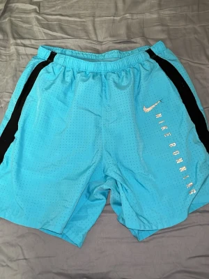 Nike Dri-Fit blå träningsshorts eller badbyxor  - Säljer ett par ljusblå Nike Dri-Fit träningsshorts eller badbyxor, med svarta detaljer längs sidorna och Nike Runner-tryck i silver. Shortsen har elastisk midja och är tillverkade i ett lätt, ventilerande syntetmaterial som passar perfekt för träning. 