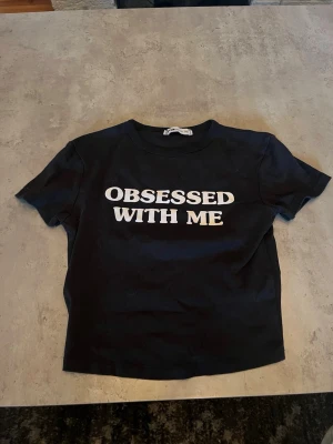 Svart t-shirt med textprint - Snygg svart t-shirt med vit text 'OBSESSED WITH ME' på bröstet. Klassisk rund halsringning och korta ärmar. Perfekt till jeans eller kjol för en cool och självsäker look.