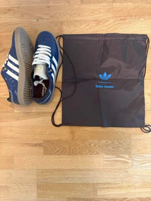 Adidas Wales Bonner blå sneakers - Snygga blå Adidas x Wales Bonner sneakers med klassiska vita ränder och gummisula. Ovandelen är i mocka och textil, med gulddetaljer på sidan och tungan. Skorna har retrovibbar och levereras med originalpåse och box. Storlek 44. MVH