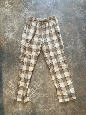 Rutiga pyjamasbyxor från Björn Borg - Säljer ett par beige och vita pyjamasbyxor från Björn Borg med rutigt mönster. Byxorna har resår i midjan och är tillverkade i ett mjukt och skönt material, perfekta för chill hemma. Klassisk och avslappnad passform.