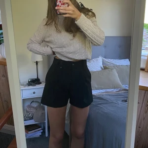 Svarta höga shorts med knapp - Snygga svarta shorts med hög midja och klassisk knappstängning. Shortsen har en stilren design med bälteshällor och raka ben. Perfekta för varmare dagar och enkla att matcha med olika toppar.