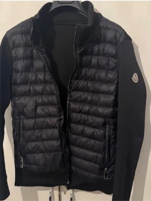 Svart pufferjacka från Moncler - Snygg svart pufferjacka från Moncler med quiltad front och släta ärmar. Jackan har hög krage, två dragkedjefickor och Moncler-logga på ena ärmen. Perfekt för kalla vinterdagar och ger en clean, modern look.