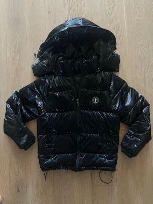 Trapstar puffer jacka  - Säljer en svart, small, glansig trapstar pufferjacka med stor huva och dragkedja framtill. Jackan har ett broderat märke på bröstet och textdetalj på ryggen. Fylld med vaddering för extra värme och har dragsko i nederkant. Perfekt för kalla vinterdagar.