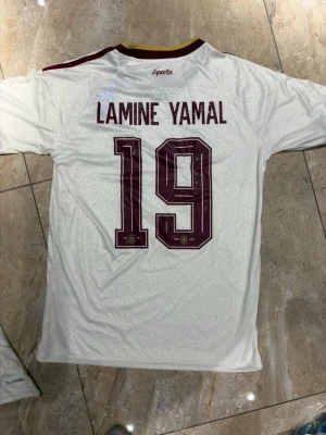 Vit Spanien fotbollströja nr 19 - Säljer en vit Spanien fotbollströja från Adidas med Lamine Yamal och nummer 19 på ryggen. Tröjan har vinröda detaljer, gula inslag vid kragen och tre klassiska Adidas-ränder på axlarna. Tillverkad i lätt och ventilerande material, perfekt för match eller träning.