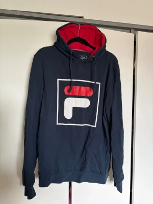 Mörkblå FILA hoodie med röd huva - Snygg mörkblå hoodie från FILA i storlek XL. Stor logga framtill och kontrastfärgad röd insida på huvan. 