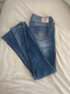 Blå jeans med röda sömmar True Religion - Säljer ett par blå jeans från True Religion med ikoniska röda kontrastsömmar och klassisk fickdesign bak. Jeansen har low midja, fem fickor och raka ben. Materialet är denim i bomull med lätt tvättad look. Perfekta för dig som gillar streetwear och snygga detaljer.