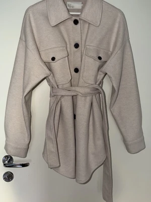 Beige kappa med knytskärp -  beige kappa med knytskärp i midjan, stora bröstfickor och svarta knappar. Overshirten har klassisk krage och långa ärmar. Perfekt för lager på lager och ger en avslappnad men trendig look.