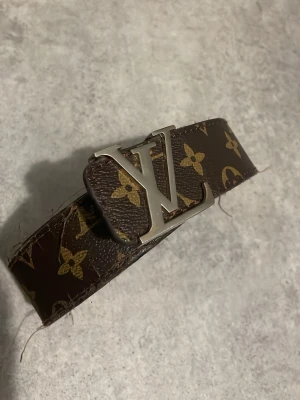 Brunt monogram skärp - Snyggt brunt skärp med klassiskt monogrammönster och stor logga i silver på spännet. Bältet är gjort i skinn och har guldfärgade detaljer i mönstret. Perfekt accessoar för att lyfta din outfit.