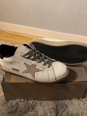 Golfen goose ÖPPEN FÖR ALLA BUD  - Snygga vita sneakers med grå stjärndetalj på sidan och grå skosnören. Skorna har svart hälparti och beige sula. Klassisk låg modell med stilren design, perfekta för en trendig vardagslook. Materialet ser ut att vara läder eller konstläder.