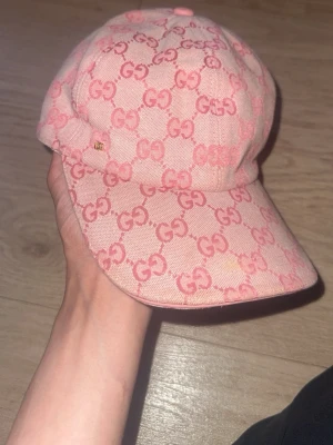 Rosa Gucci keps med GG-mönster - Säljer en snygg rosa keps från Gucci med det klassiska GG-mönstret i en ljus och mörkare rosa ton. Kepsen har böjd skärm och justerbar rem bak. Tillverkad i canvasmaterial med insida i bomull. Perfekt för dig som vill ha en trendig och lyxig accessoar.