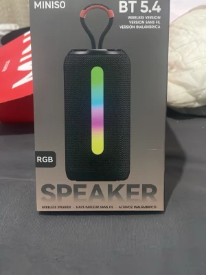 Miniso bärbar högtalare med RGB-ljus - Snygg bärbar högtalare från Miniso med RGB-ljus i mitten. Högtalaren är svart med cylinderform och har ett handtag upptill i orange. Trådlös via Bluetooth 5.4 och tillverkad i plast och tyg. Perfekt för att ta med sig överallt.
