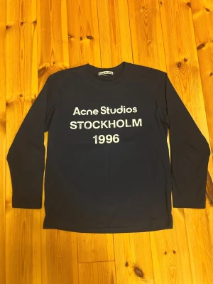 Mörkblå långärmad t-shirt Acne Studios - Snygg mörkblå långärmad tröja från Acne Studios med den klassiska vita texten “Acne Studios STOCKHOLM 1996” på bröstet. Tröjan har rund halsringning och en stilren passform som passar perfekt till både vardag och finare tillfällen.  Använd endast ett fåtal gånger och är i mycket fint skick. Säljs på grund av att den tyvärr är för liten.
